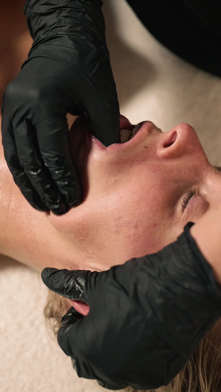Behandlerens hænder med handsker masserer indersiden af kinden under Buccal Massage.